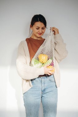 Beyaz güneşli bir odada elinde sebze ağıyla poz veren mutlu bir kadın.