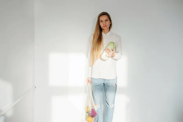 Mujer sin emociones en una habitación blanca soleada posando, sosteniendo la bolsa de red con verduras —  Fotos de Stock