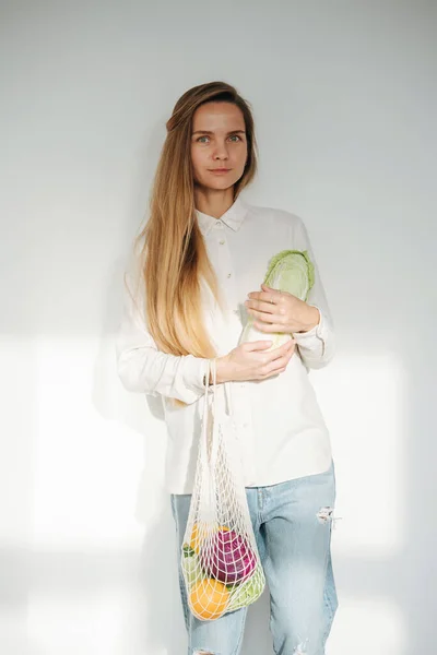 Subtely smiling modest woman posing in a room, holding net bag with vegetables