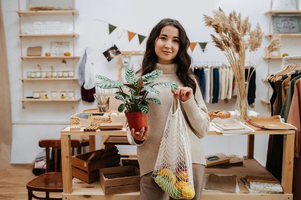 Портрет женщины в эко-магазине, позирующей для фото с сетчатой сумкой и цветочным горшком — стоковое фото