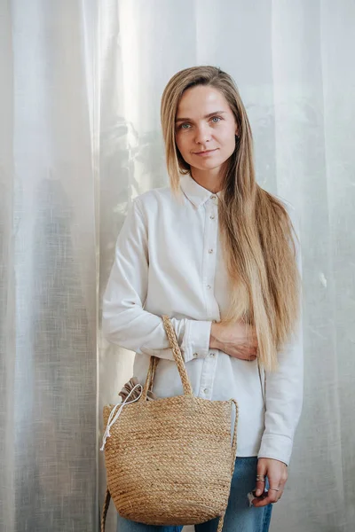 Modest woman in white sunlit room posing for a photo, holding wicker bag