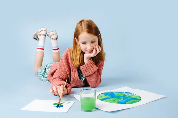 Chica pelirroja tirada en el suelo dibuja un planeta verde-azul —  Fotos de Stock