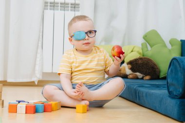 Little boy in lazy eye patch eating apple on the children room clipart