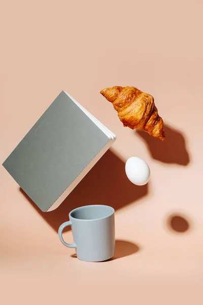 Taza de café en la superficie de color melocotón. Croissant, huevo y cuaderno flotando en el aire — Foto de Stock