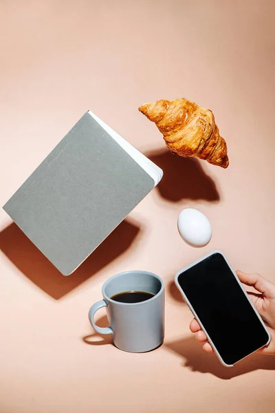 Hand taking picture on a phone. Croissant, egg and notebook hovering in air — Stock Photo, Image