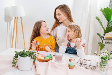 Anne iki kızıyla vakit geçiriyor, masanın arkasına paskalya yumurtası çiziyor.