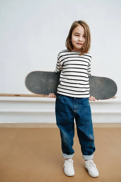 Маленькая девочка позирует со скейтбордом для фото, фальшивая уставшая улыбка — стоковое фото