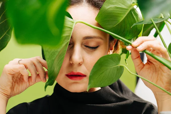 Sensual mulher de meia idade com os olhos fechados atrás de grandes folhas de plantas. — Fotografia de Stock