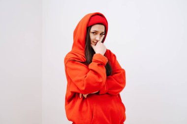 Kırmızı kapüşonlu kazaklı ve şapkalı genç bir kız elini yüzünden tutuyor.