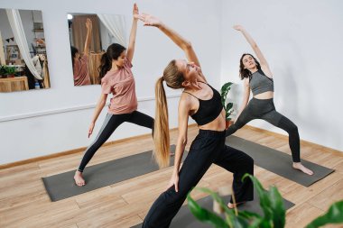 Eğitmen önünde yoga yapan esnek kadınlar, ellerinde savaşçı pozuyla.