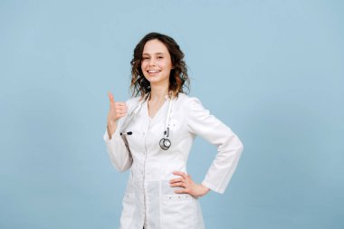 Parlak gülümseyen kadın hemşire baş parmağını kaldırıyor. Mavi arka planda. Beyaz bornoz ve steteskop giyiyor..