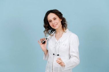 Gülümseyen ve konuşan arkadaş canlısı bir hemşire. Mavi arka planda. Beyaz bornoz ve steteskop giyiyor..