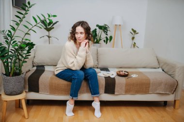Düşünceli genç bir kadın dairesinin konforlu koltuğunda nakış aletleri ve malzemelerinin yanında oturuyor..