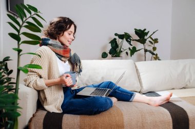 Sıcak giyimli genç bir bayan evinin konforunda dizüstü bilgisayar izliyor. Kanepede oturuyor, atkı takıyor, kahve içiyor. Yan görünüm.