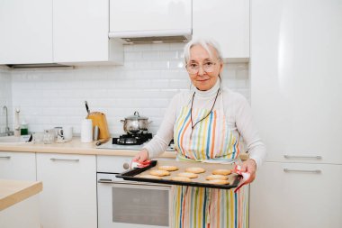 Mutfakta duran önlüklü, yaşlı, kır saçlı bir kadın fırından kurabiyeleri çıkardı.. 