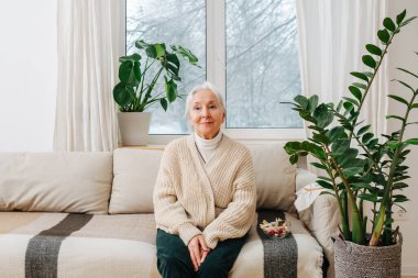 Evde koltukta oturan, elleri dizlerinin arasında gülümseyen pozitif bir büyükanne. Arkasında kış manzaralı bir pencere var..