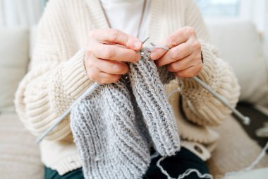 Büyükanne evde bir kanepede oturuyor, iğnelerle örgü örüyor, gri yün kullanıyor. Kırpılmış, yüzü yok.