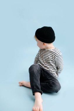 Bacak bacak üstüne atmış yerde oturan küçük sarışın çocuk. Shoukder 'ının arkasına bakıyor. Çizgili uzun kollu gömlek ve siyah şapka giyiyor. Stüdyo çekimi. Mavi arkaplan.