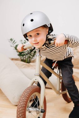 Sevimli küçük sarışın çocuk bir odada bej renkli iki tekerlekli bisiklette oturuyor.