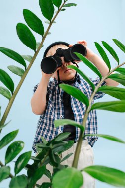 Dürbünle bakan, bitki dallarının arkasında duran meraklı sarışın çocuk.