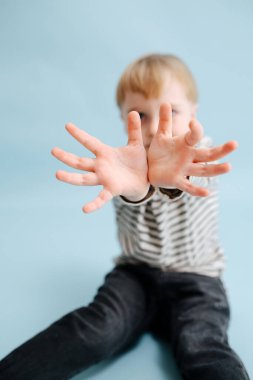 Sarışın çocuk yerde oturuyor, bileklerini kameraya yakın tutuyor. Seçici olarak ellere odaklan. Çizgili uzun kollu gömlek giyiyor. Stüdyo çekimi. Mavi arkaplan.