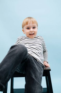 İskemlede oturan şakacı sarışın çocuk. Çizgili uzun kollu gömlek giyiyor. Stüdyo çekimi. Mavi arka planda. Bacağıyla kameraya uzanıyor..