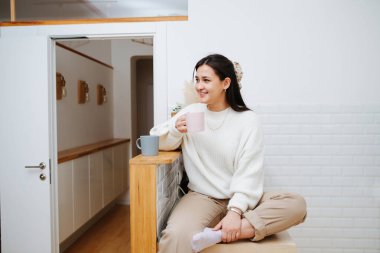 Güzel genç bir kadın bacağı yukarıda, yüksek bir kithen masasında oturuyor, bir kupa şeklinde içiyor. Gülümsüyor ve birine bakıyor. Sıradan giyinmiş..