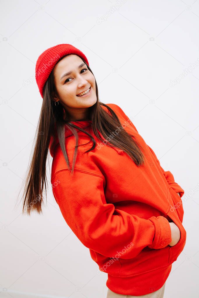 Engaiged smilig woman in vibrant red cap and hoodie tilting back with hands in pockets. She is standing sideways, looking at the camera. Indoors, in front of the white wall.