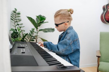 Güneş gözlüklü havalı çocuk evde elektrikli piyano çalıyor. Kot pantolon ceketi giyiyor, sarı saçları topuz şeklinde. Yan görünüm.