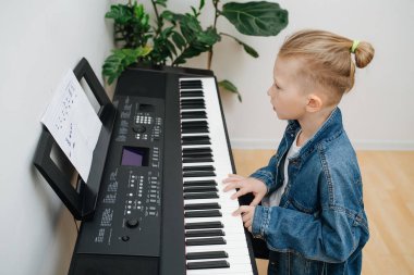 Odaklanmış küçük çocuk evde elektrikli piyano çalmayı öğreniyor. O, bir kalemle yazdığı notaları okuyor. Kot pantolon ceketi giyiyor, sarı saçları topuz şeklinde. Yüksek açı