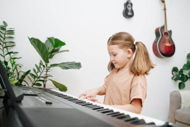 Sevimli küçük kız evde elektrikli piyano çalmaktan hoşlanıyor. Yan görüş. Saçları iki kalın at kuyruğu şeklinde. Sadece bir kaç parmağını kullanıyor. Yan görünüm.