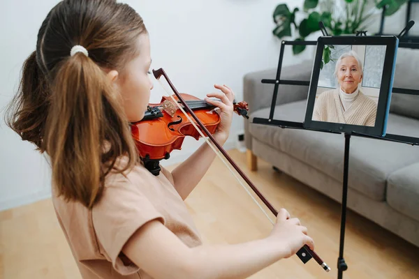 Little girl with two pony tails learning to play violin, showing her progress to her grandma in a video chat. The tablet on a music stand.