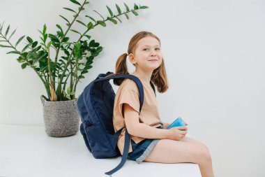 Küçük liseli kız masada oturuyor, birine gülümsüyor. Sırtında sırt çantası var, yemeğini bir kutuda tutuyor, okula gitmeye hazır. Beyaz arkaplan.