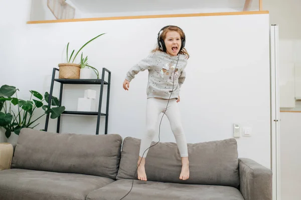 精力充沛的小女孩在沙发上跳着 一边用大耳机听着精力充沛的音乐 一边没人在看 在客厅里 感觉冲动 做面部表情 — 图库照片