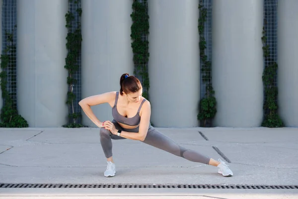 Keen woman stretching on a concrete paving, doing side lounges. Over support pillars. In grey top and yoga pants. She's consentrated on tension in her muscles.