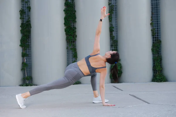 Strong woman doing yoga on a concrete paving, doing warrior asana with a twist. Over support pillars. In grey top and yoga pants. Side view.