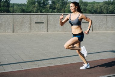 Exhilarated athletic woman running fast outdoors on the track next to pavement sidewalk. On a sunny day under a clear blue sky. She is wearing grey top and mini shorts. clipart