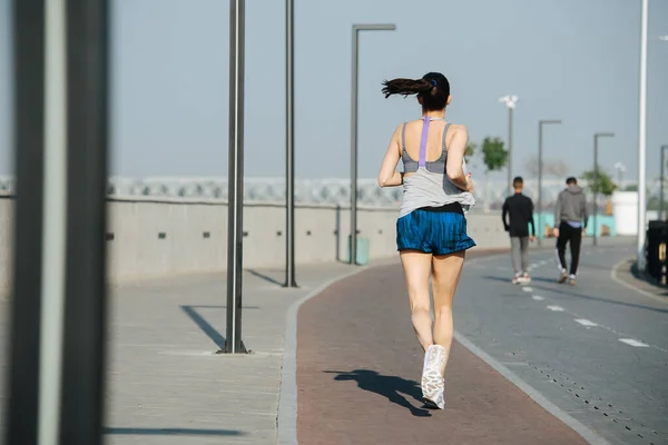 歩道の横の新しいトラックの屋外で逃げ背の高い運動選手の女性 晴れた日には青空の下で 彼女は白い袖のないシャツとミニショートパンツを着ています — ストック写真