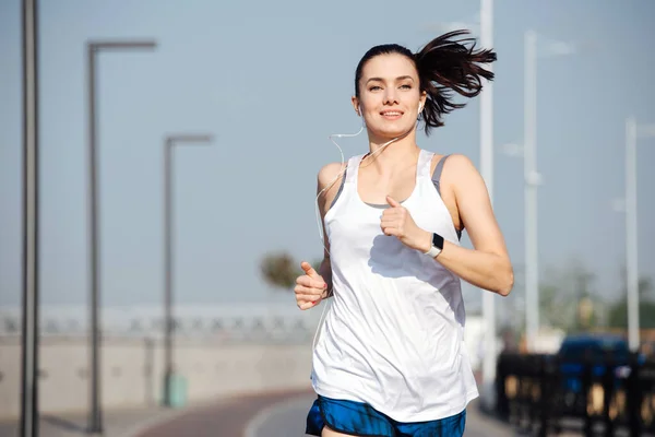 Açık havada koşan kendinden geçmiş atletik bir kadın. Arka plan bulanık. Açık mavi gökyüzünün altında güneşli bir günde. Beyaz kolsuz gömlek ve mini şort giyiyor..