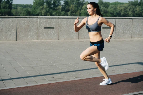 Exhilarated athletic woman running fast outdoors on the track next to pavement sidewalk. On a sunny day under a clear blue sky. She is wearing grey top and mini shorts.