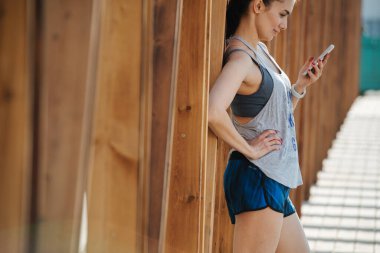 Kendine güvenen seksi esmer açık havada telefonla mesajlaşıyor, ahşap çerçeveye yaslanıyor.