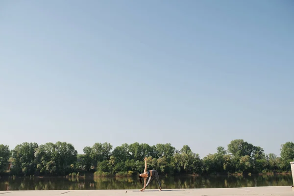 Långt skott av en kvinna som gör yoga på trottoaren på en flod — Stockfoto