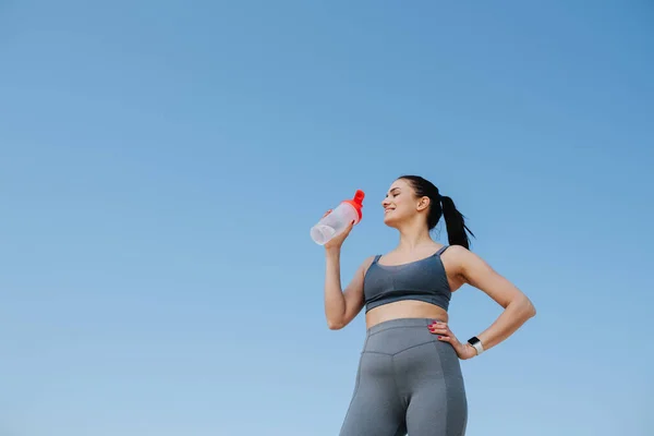 Улыбающаяся молодая женщина в спортивной одежде пьет воду. На голубом небе — стоковое фото