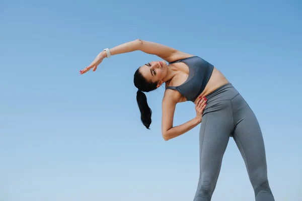 Mooie vrouw in sportkleding die zich uitstrekt. Boven de blauwe lucht. Opwaartse hoek — Stockfoto