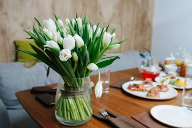 Sıcak bir yemek masasında vazoda duran bir buket taze lale çiçeği. Bulanık arkaplandaki abur cubur çeşitliliği.