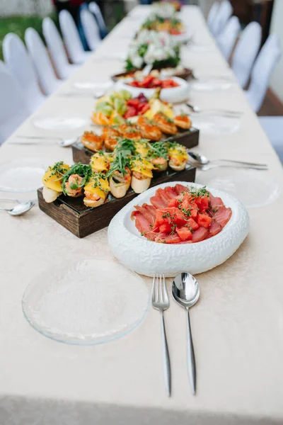 Narrow long dining table outdoors, abundant in the food. Dishes layed in a straight line on a thick wooden boards and plates.