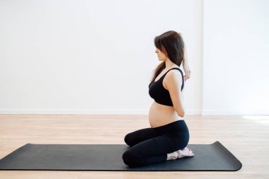 Hamile bir kadının dizlerinin üstünde sırtüstü oturup büyük bir stüdyoda yoga çalışmasının yan görüntüsü. Arkasından parmaklarını tutuyor. Siyah bluz ve pantolon giyiyor.