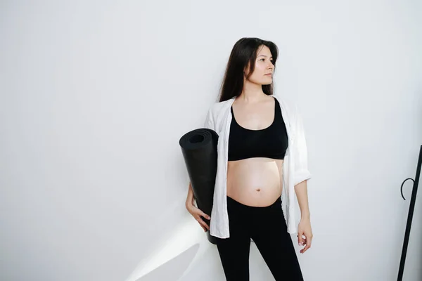 ヨガマットを手にした妊婦の肖像画 黒のトップスとパンツを着てる 横を見ると — ストック写真