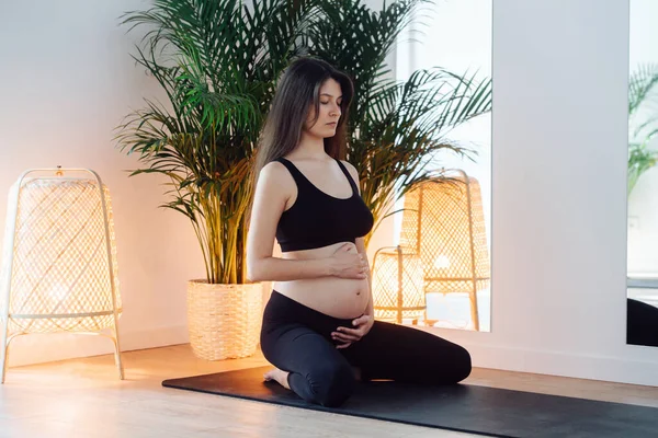 Calm pregnant woman sitting on her knees, doing breathing exercises. She\'s wearing black top and pants. She\'s meditating, holding her hands on a belly. Intimate illumination in a yoga studio.