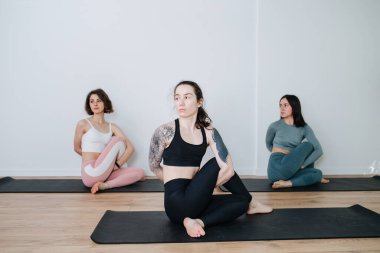 Spor kıyafetleriyle yoga yapan sakin, biraz sıkılmış kadınlar büyük bir stüdyoda yan yana esneme hareketleri yapıyorlar. Büyük omuzlu dövmeli bir kıza odaklan.